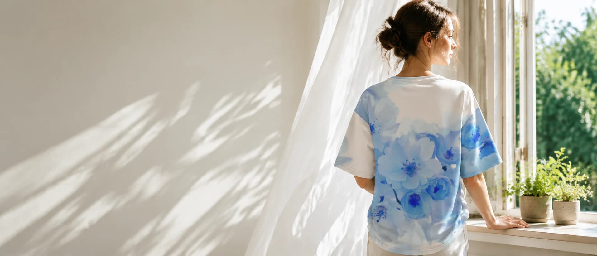 Woman looking out a window wearing a white t-shirt with a soft blue floral print.